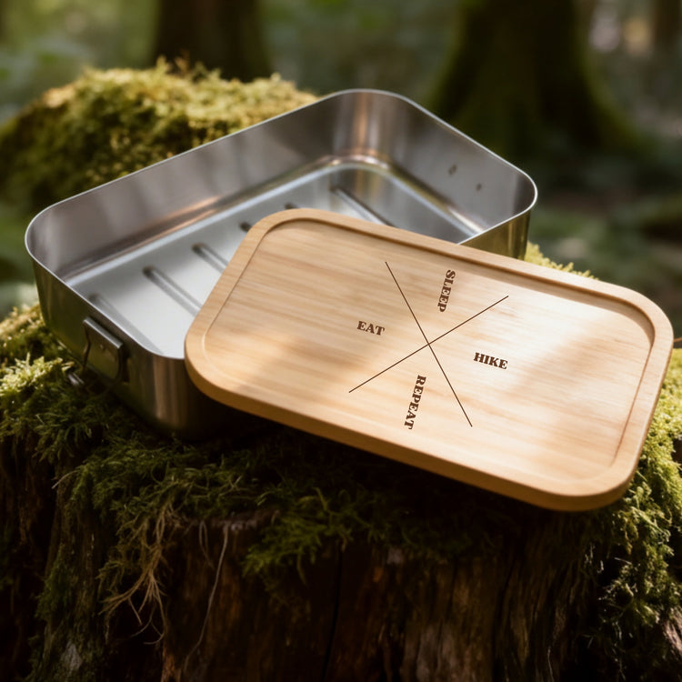 Edelstahl-Brotdose mit Bambusdeckel und Gravur Eat Sleep Hike Repeat auf moosigem Baumstumpf im Wald.