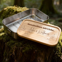 Personalisierte Edelstahl-Brotdose mit Bambusdeckel und graviertem Schriftzug auf moosbedecktem Baumstamm im Wald.