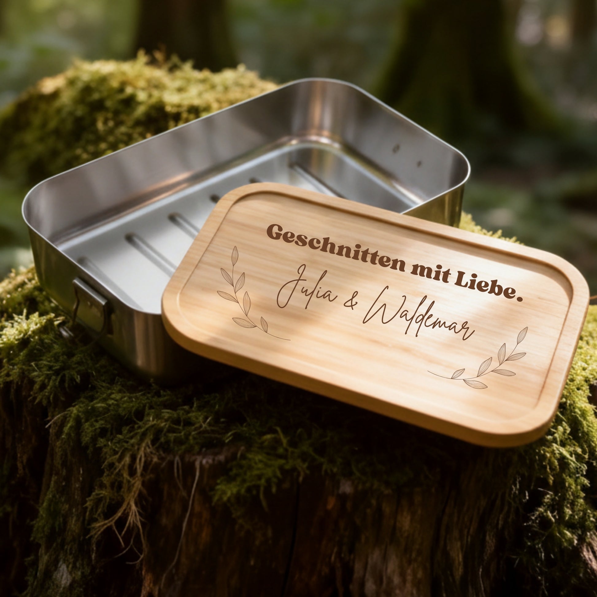 Personalisierte Edelstahl-Brotdose mit Bambusdeckel und graviertem Schriftzug auf moosbedecktem Baumstamm im Wald.
