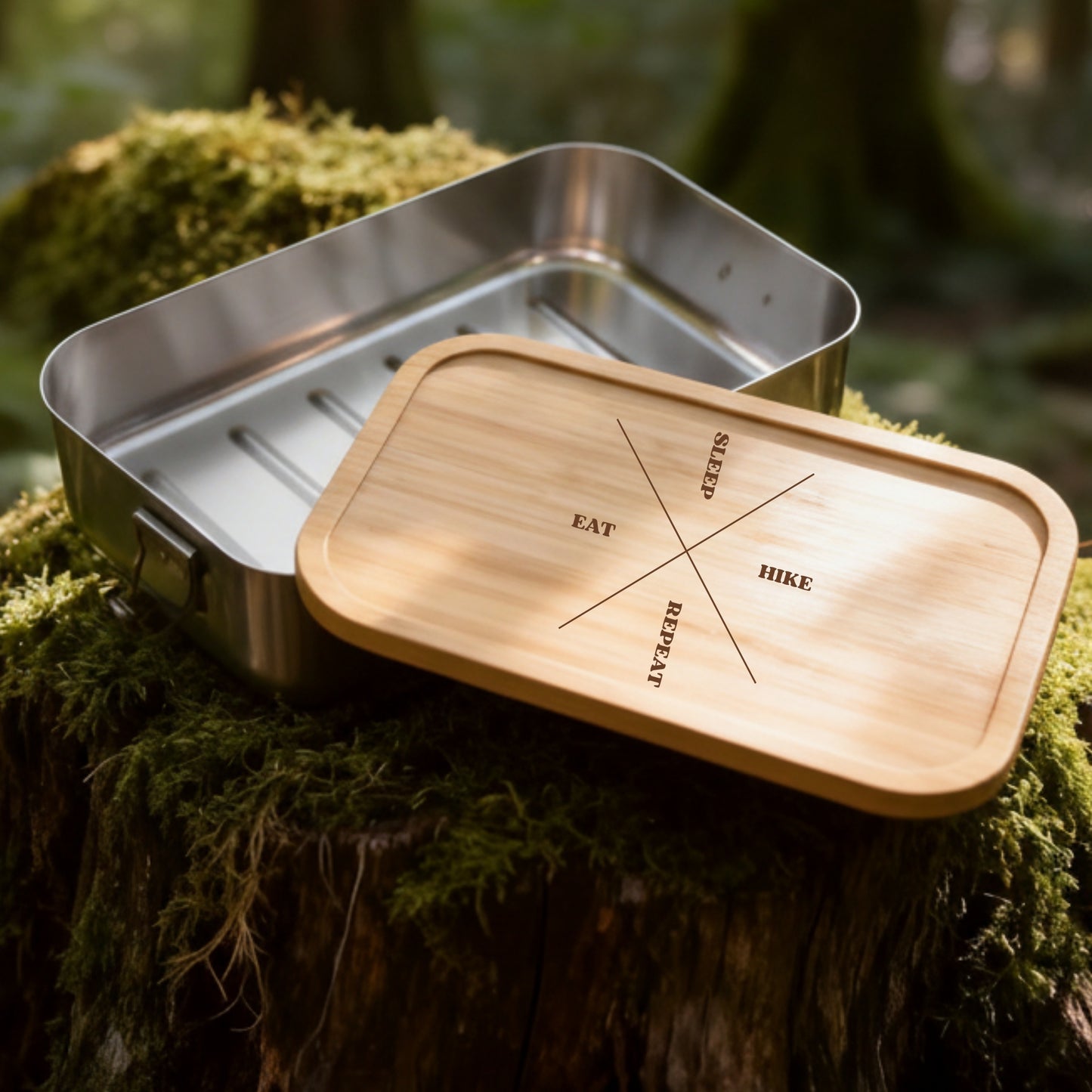 Edelstahl-Brotdose mit Bambusdeckel und Gravur Eat Sleep Hike Repeat auf moosigem Baumstumpf im Wald.