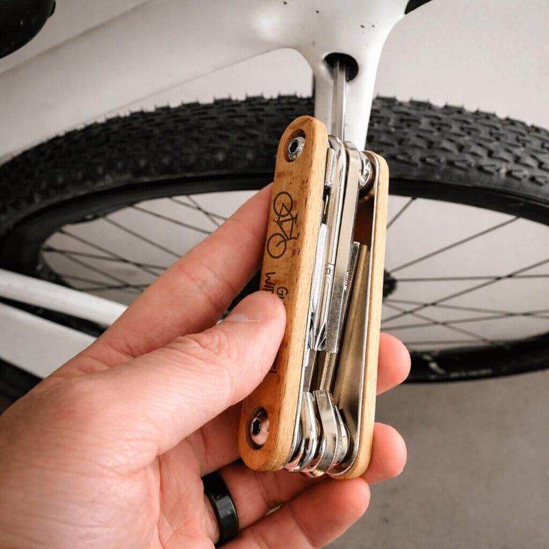 Fahrrad Multitool 9in1 aus Edelstahl mit graviertem Holzgriff und Fahrradsymbol, gehalten vor einem Rad.