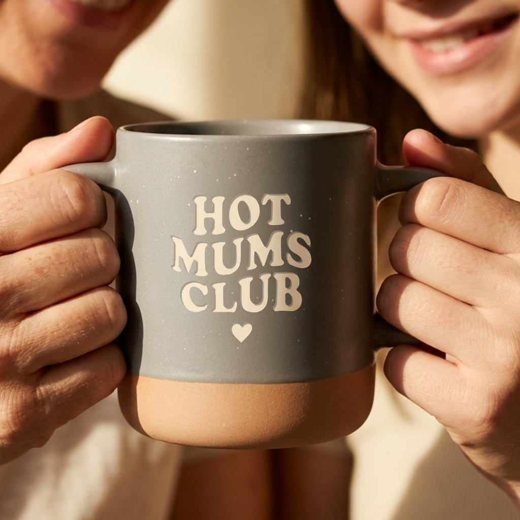 Graue Keramiktasse mit Hot Mums Club Aufdruck und Herz, von zwei Frauen gehalten.
