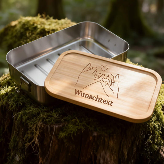 Edelstahl-Brotdose mit graviertem Bambusdeckel und personalisiertem Wunschtext auf moosigem Baumstumpf im Wald.
