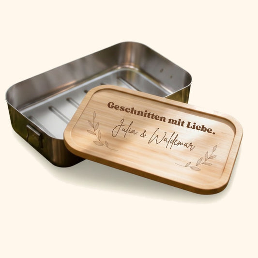 Personalisierte Edelstahl-Brotdose mit Bambusdeckel und graviertem Schriftzug auf moosbedecktem Baumstamm im Wald. — vor cremefarbenem Hintergrund.