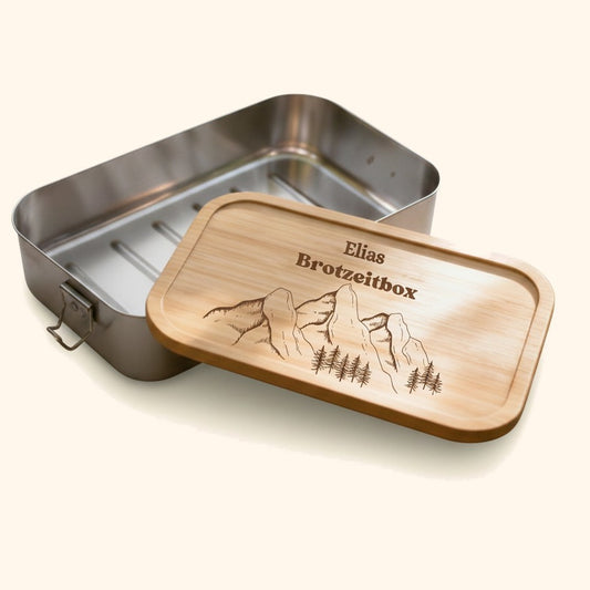 Personalisierte Edelstahl-Brotdose mit Bambusdeckel und graviertem Bergmotiv samt Name auf moosigem Baumstumpf. — vor cremefarbenem Hintergrund.