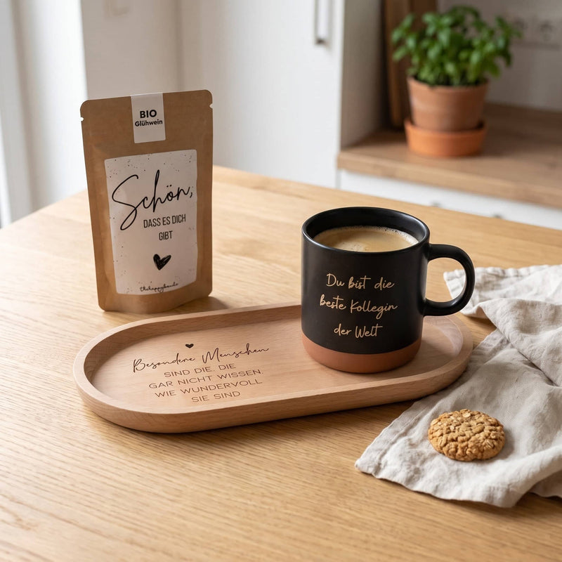 Geschenkset Kaffeepause auf einem Küchentisch: gravierte schwarze Tasse mit Kaffee auf dem Holz-Tablett, daneben das Sprüche-Tütchen und ein Keks.