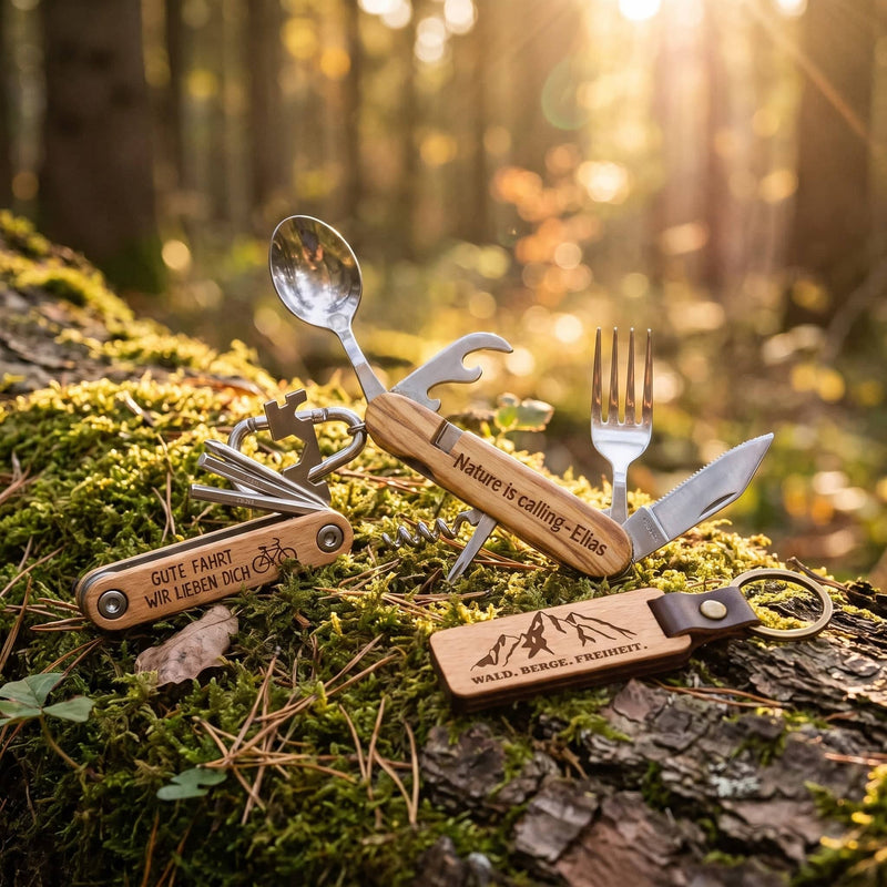 Campingbesteck, Fahrrad-Multitool und Schlüsselanhänger „Wald. Berge. Freiheit.“ auf einem moosbewachsenen Baumstamm im Abendlicht.