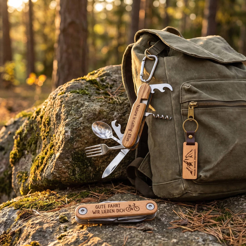 3er Set Outdoor am Wanderrucksack im Wald: Campingbesteck am Karabiner, Schlüsselanhänger am Reißverschluss, Fahrrad-Multitool auf dem Felsen davor.