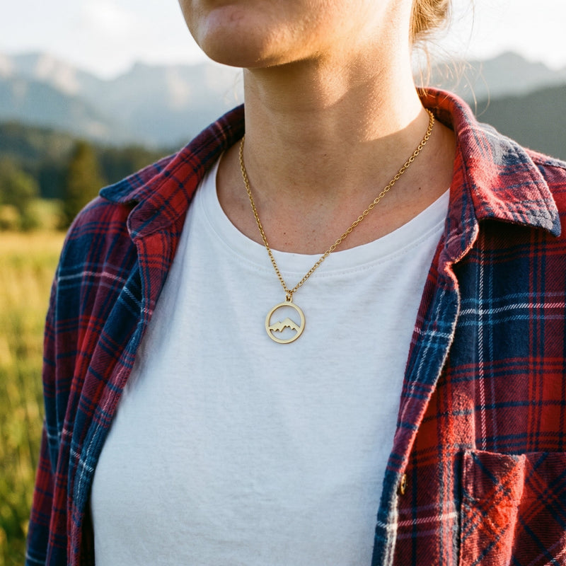 Frau trägt die goldfarbene Halskette mit Berg-Anhänger aus Edelstahl über einem weißen T-Shirt in den Bergen.