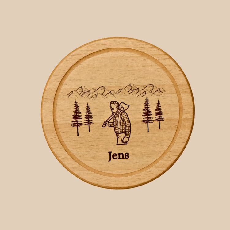 Rundes Holzbrett aus Buche mit personalisierter Gravur eines Holzfällers, Bergpanorama und dem Namen Jens.