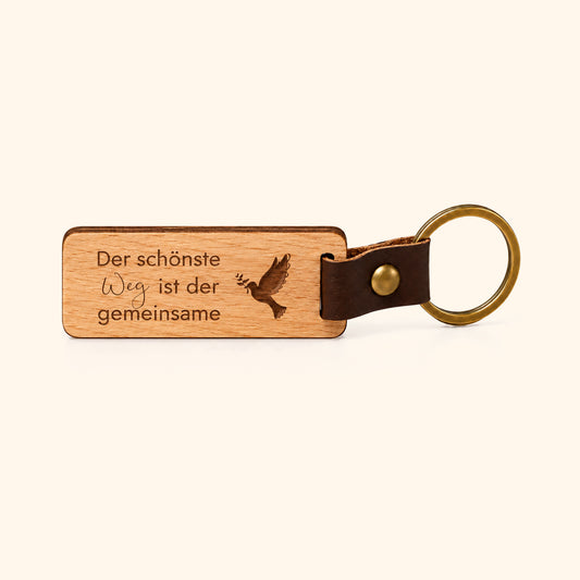 Schlüsselanhänger aus Buchenholz mit Lederlasche und Messingring, Gravur „Der schönste Weg ist der gemeinsame“ mit Taube, vor cremefarbenem Hintergrund.