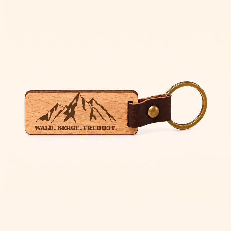 Gratis im Set: Schlüsselanhänger aus Buche mit Bergmotiv und „Wald. Berge. Freiheit.“ vor cremefarbenem Hintergrund.