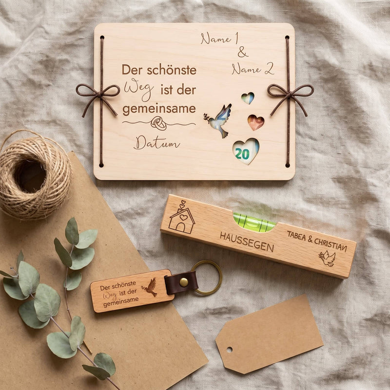 3er Set aus gravierter Geldgeschenk-Box, Haussegen Wasserwaage und Schlüsselanhänger von oben auf Leinen, daneben Kraftpapier, Juteschnur und Eukalyptus.