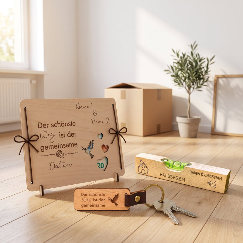 Personalisierte Geldgeschenk-Box, Haussegen Wasserwaage und Schlüsselanhänger mit Haustürschlüsseln im neuen Zuhause neben Umzugskarton und Olivenbaum.