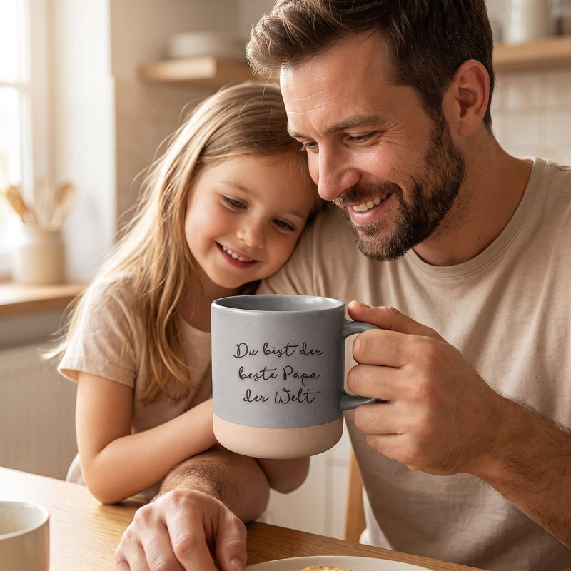 Graue Keramiktasse mit gravierter Handschrift „Du bist der beste Papa der Welt“, Vater und Tochter halten sie gemeinsam am Frühstückstisch.