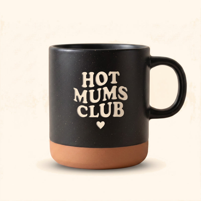 Schwarze Keramiktasse mit personalisiertem Druck "Hot Mums Club" und Herz als Muttertagsgeschenk. — vor cremefarbenem Hintergrund.