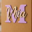 Personalisiertes Holz-Türschild in Flieder mit geschwungenem Namenszug an heller Zimmertür.