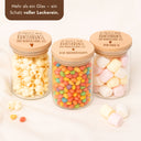 Drei Vorratsgläser mit graviertem Holzdeckel und Danke-Schriftzug, gefüllt mit Popcorn, bunten Bonbons und Marshmallows.
