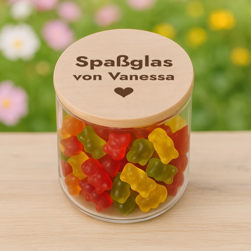 Personalisiertes Vorratsglas mit Holzdeckel-Gravur „Spaßglas von Vanessa“ und Herz auf einem Holztisch im Garten, gefüllt mit Gummibärchen.