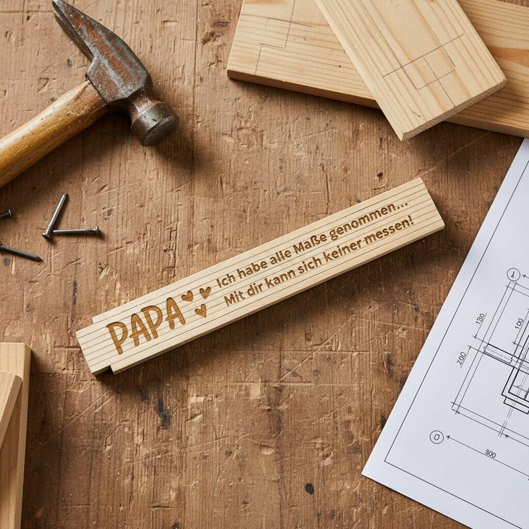 Personalisierter Zollstock aus Holz mit Gravur Papa auf einer Werkbank neben Hammer und Bauplan.