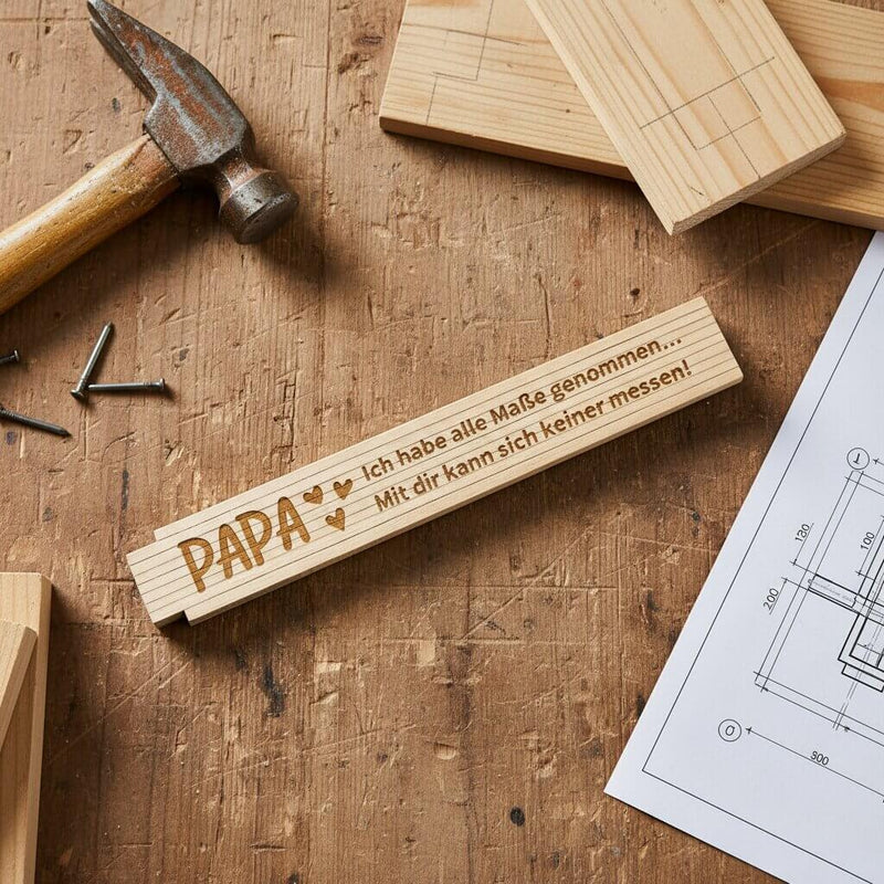 Personalisierter Zollstock aus Holz mit Gravur Papa auf einer Werkbank neben Hammer und Bauplan.