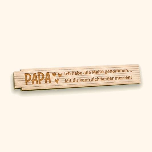 Personalisierter Zollstock aus Holz mit Gravur Papa-Spruch als Geschenkidee zum Vatertag. — vor cremefarbenem Hintergrund.